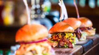 Close up image of a selection of freshly flame grilled burgers in a row on a wooden counter at Borough Market, one of the oldest and most famous food markets in the world. Each of the burgers has its own label, on which is written the contents of the burger. The burgers are sandwiched between glazed buns, and presented on beds of fresh green lettuce and stuffed with melted cheese and red onion. Horizontal colour image with copy space.