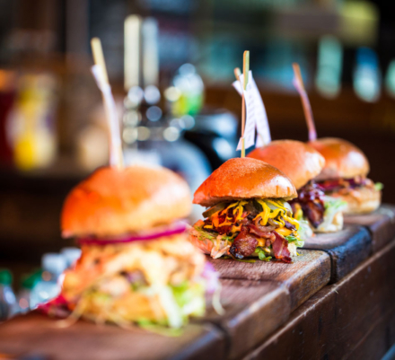 Close up image of a selection of freshly flame grilled burgers in a row on a wooden counter at Borough Market, one of the oldest and most famous food markets in the world. Each of the burgers has its own label, on which is written the contents of the burger. The burgers are sandwiched between glazed buns, and presented on beds of fresh green lettuce and stuffed with melted cheese and red onion. Horizontal colour image with copy space.